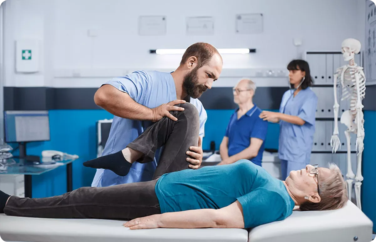 A physiotherapist guiding an older patient through rehabilitation and leg stretching exercises while medical staff care for other patients in the background, a private polyclinic in Belgrade.